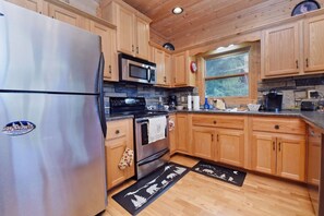 A kitchen with wooden cabinets, a stainless steel refrigerator, a microwave, a stove, and a window with a view of greenery. - Work Hard Play Hard- View-Gameroom (Blue Ridge)