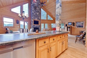 A spacious kitchen with a stone fireplace, wooden cabinets, and a large island with a granite countertop. - Work Hard Play Hard- View-Gameroom (Blue Ridge)