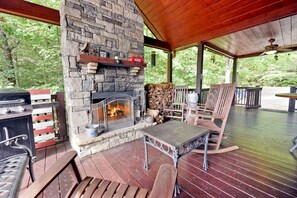 A stone fireplace with a fire, surrounded by wooden furniture and a grill on a covered deck. - Work Hard Play Hard- View-Gameroom (Blue Ridge)
