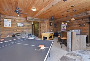 Une table de ping-pong dans une pièce aux murs et au plafond en bois, une table de baby-foot et un coin bar avec des bouteilles.