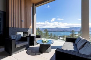 A modern outdoor seating area with wicker furniture, a round table with a floral centerpiece, and a view of a lake and mountains.