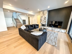 A modern living room with a dark sofa, a coffee table, and a fireplace. A staircase is visible in the background.