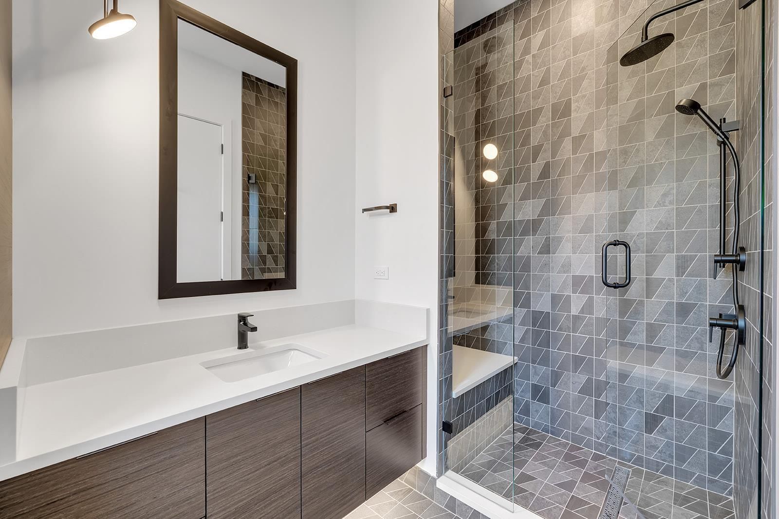 Ein modernes Badezimmer mit einem weißen Waschbecken, dunklen Holzschränken, einem großen Spiegel und einer Dusche mit einem geometrischen Fliesenmuster.