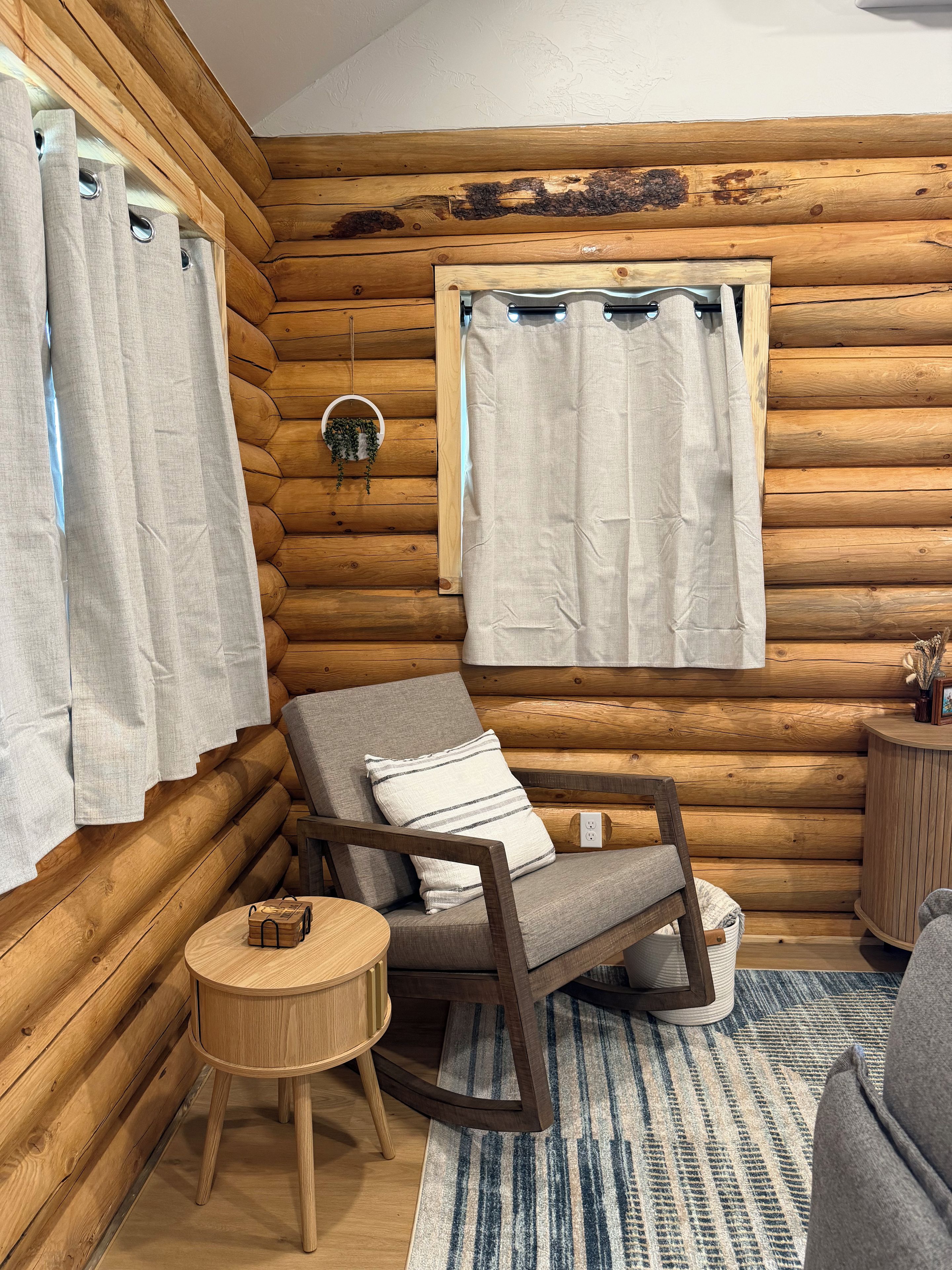 Interior de una cabaña de troncos con una silla de madera, una mesita redonda y una ventana con cortinas.