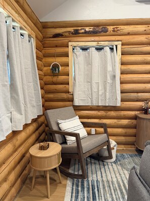 A log cabin interior with a wooden chair, a small round table, and a window with curtains.