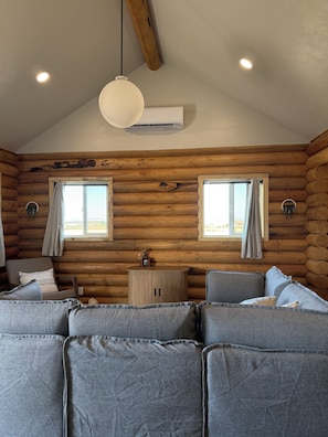 Et interiør af en hytte med en sofasæt, to vinduer med gardiner, en hængende lampe og et lille bord med en pynteting.
