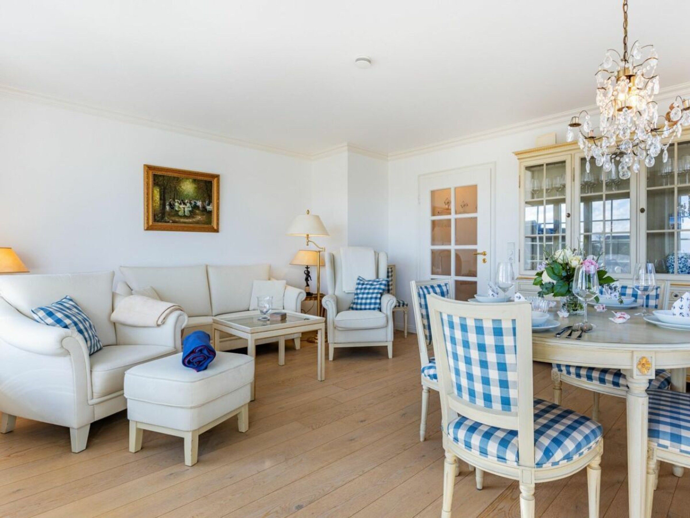 A room with a white sofa set, a dining table with checkered chairs, a chandelier, and a painting on the wall.