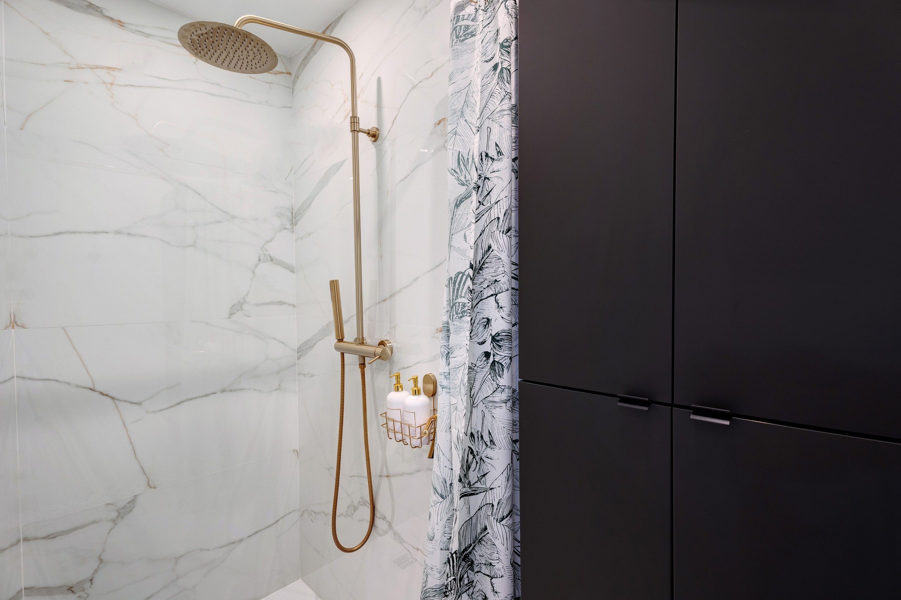 Un baño con ducha, paredes de mármol, un mueble oscuro y cortina de baño con estampado de hojas.