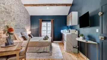 A bedroom with a stone wall, a bed with pillows, a bedside table with a vase, and a view of a kitchen with a TV.