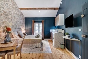 A bedroom with a stone wall, a bed with pillows, a bedside table with a vase, and a view of a kitchen with a TV.