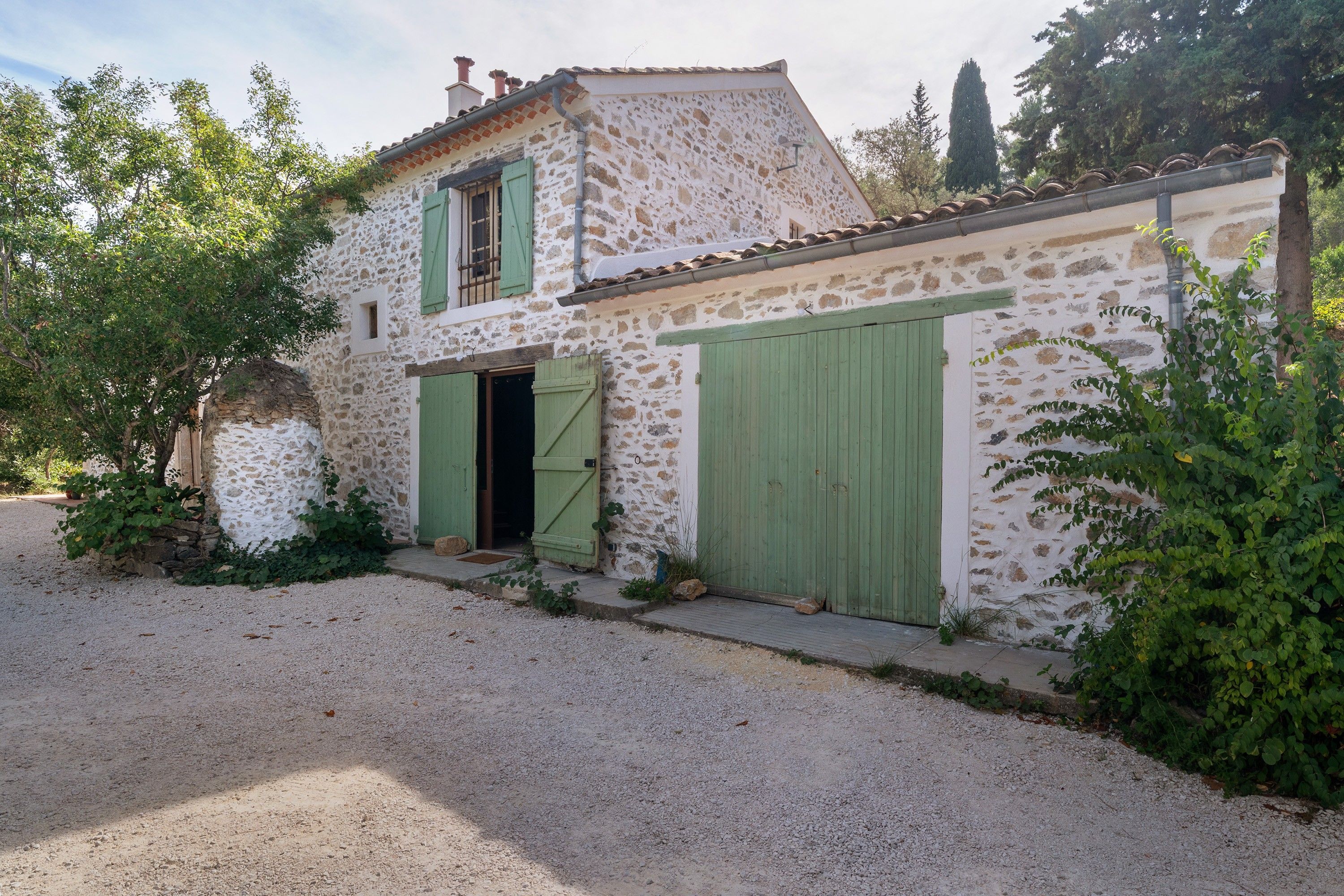 Une maison en pierre à deux étages, avec une allée en gravier et des volets verts.