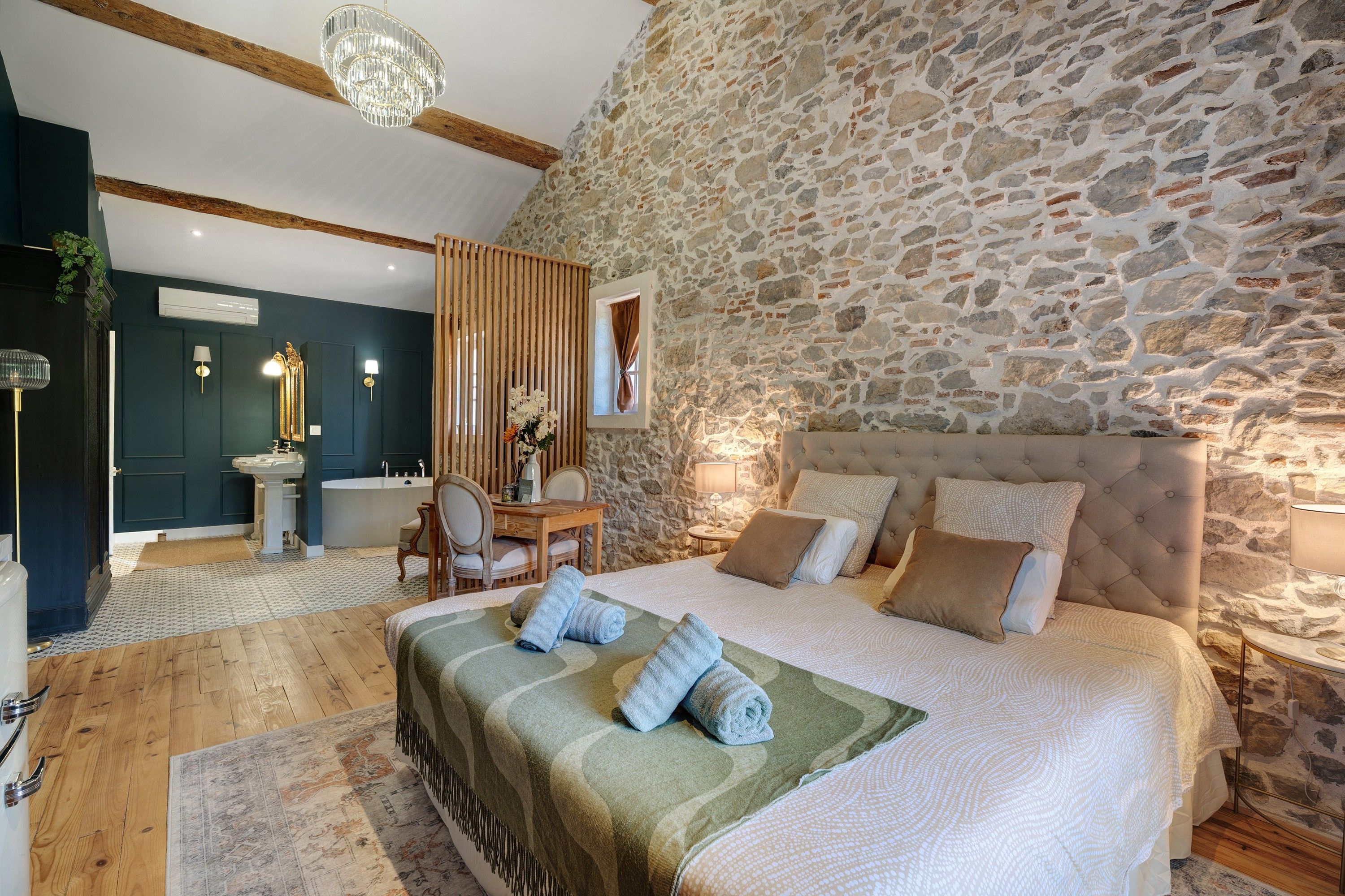 A bedroom with a stone wall, a large bed with multiple pillows, a wooden floor, and a bathroom with a bathtub and sink.