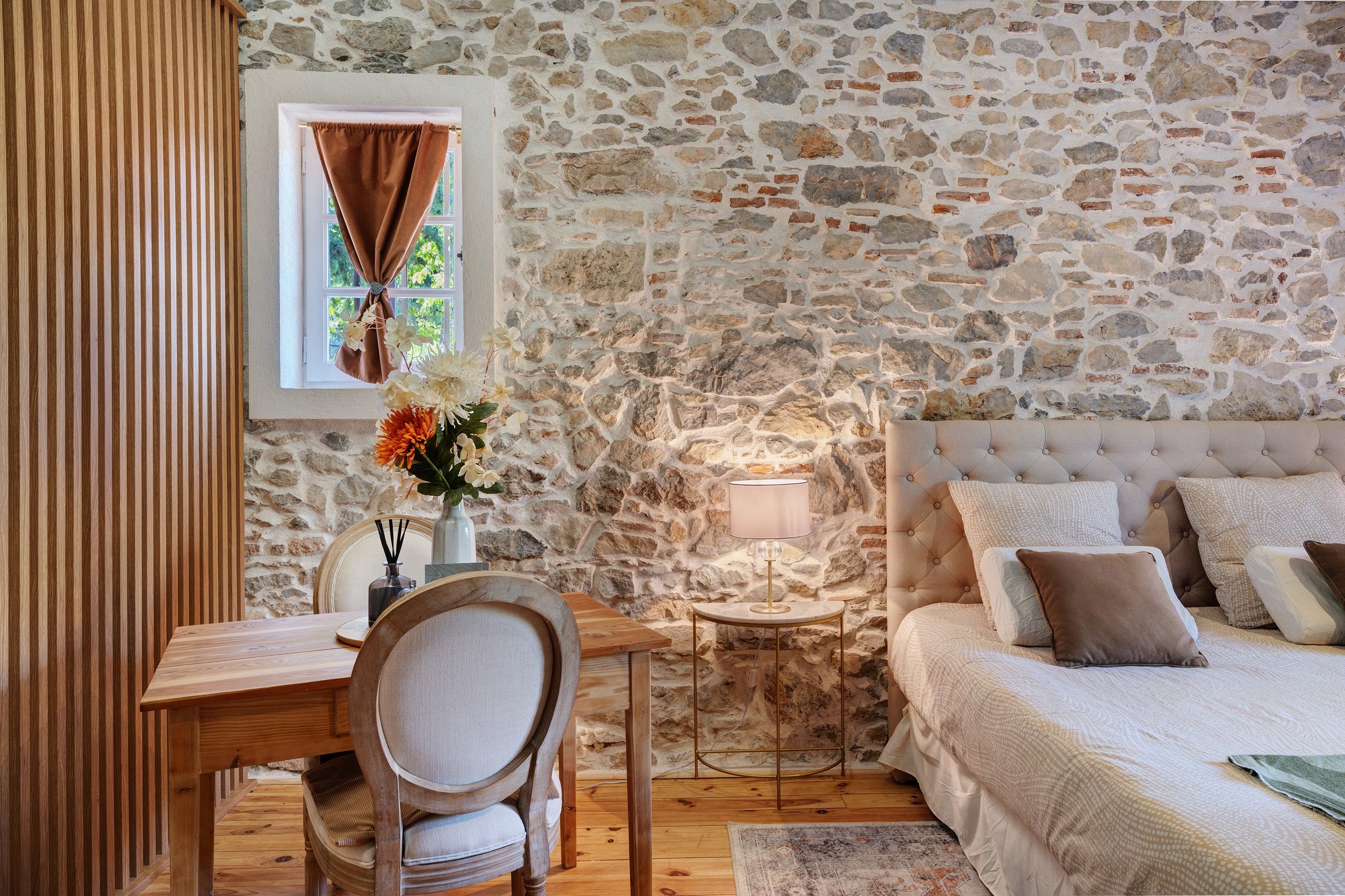 A bedroom with a stone wall, a bed with multiple pillows, a bedside table with a lamp, and a chair.