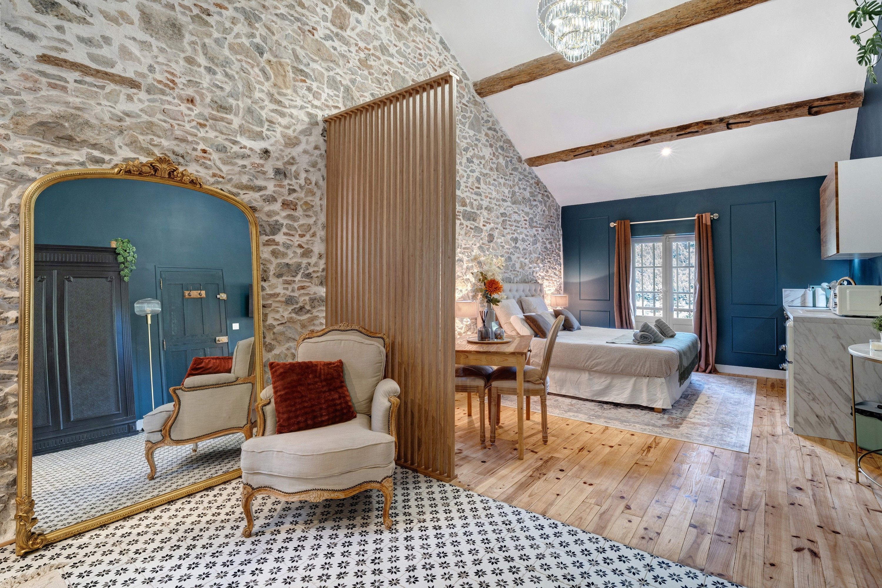 Una habitación con pared de piedra, un espejo con marco dorado, una silla de madera, una cama y una mesa de comedor con sillas.