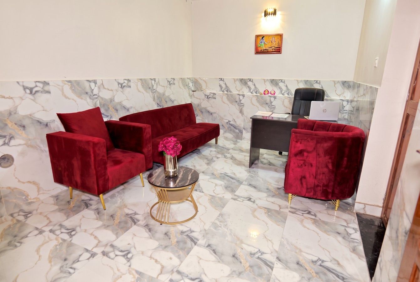 A lobby with marble flooring, red velvet sofas, a small round table with a vase of flowers, and a desk with a laptop.