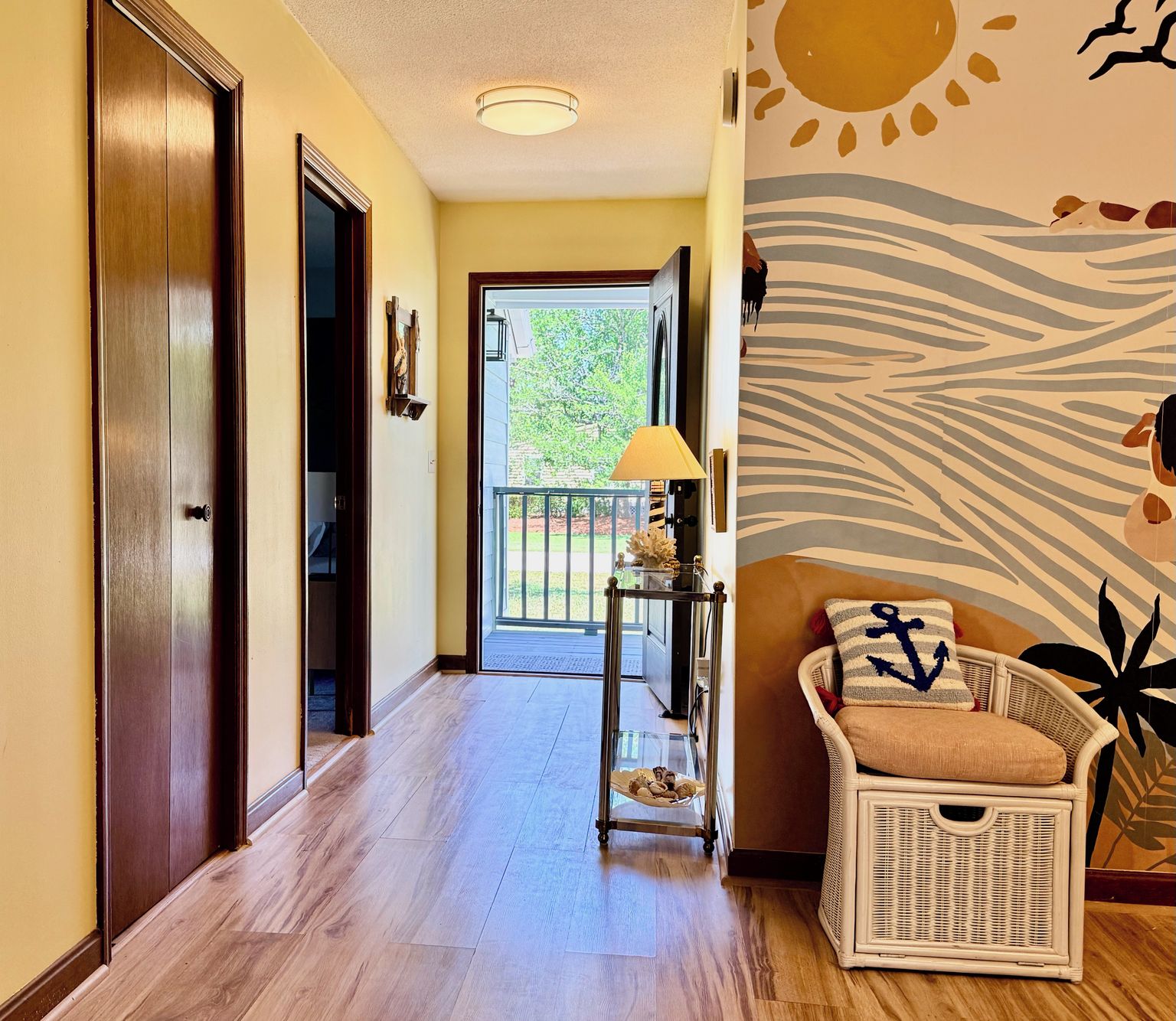A hallway with a wooden floor, a wicker chair with a cushion, a lamp, and a wall with a sun and wave design.