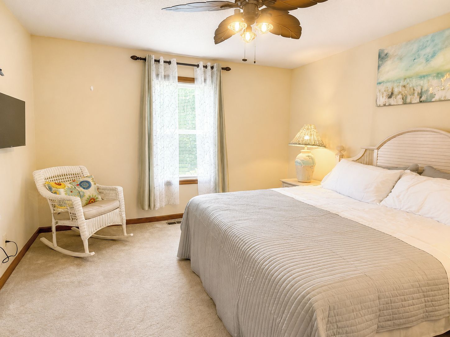 Un dormitorio con cama, una mecedora, ventilador de techo, ventana con cortinas y un cuadro en la pared.
