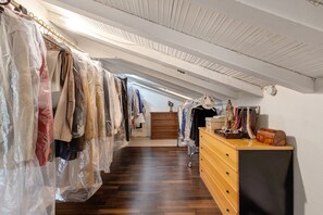 Een smalle gang met houten vloer, een houten dressoir met lades en een kledingrek met spullen in plastic zakken.