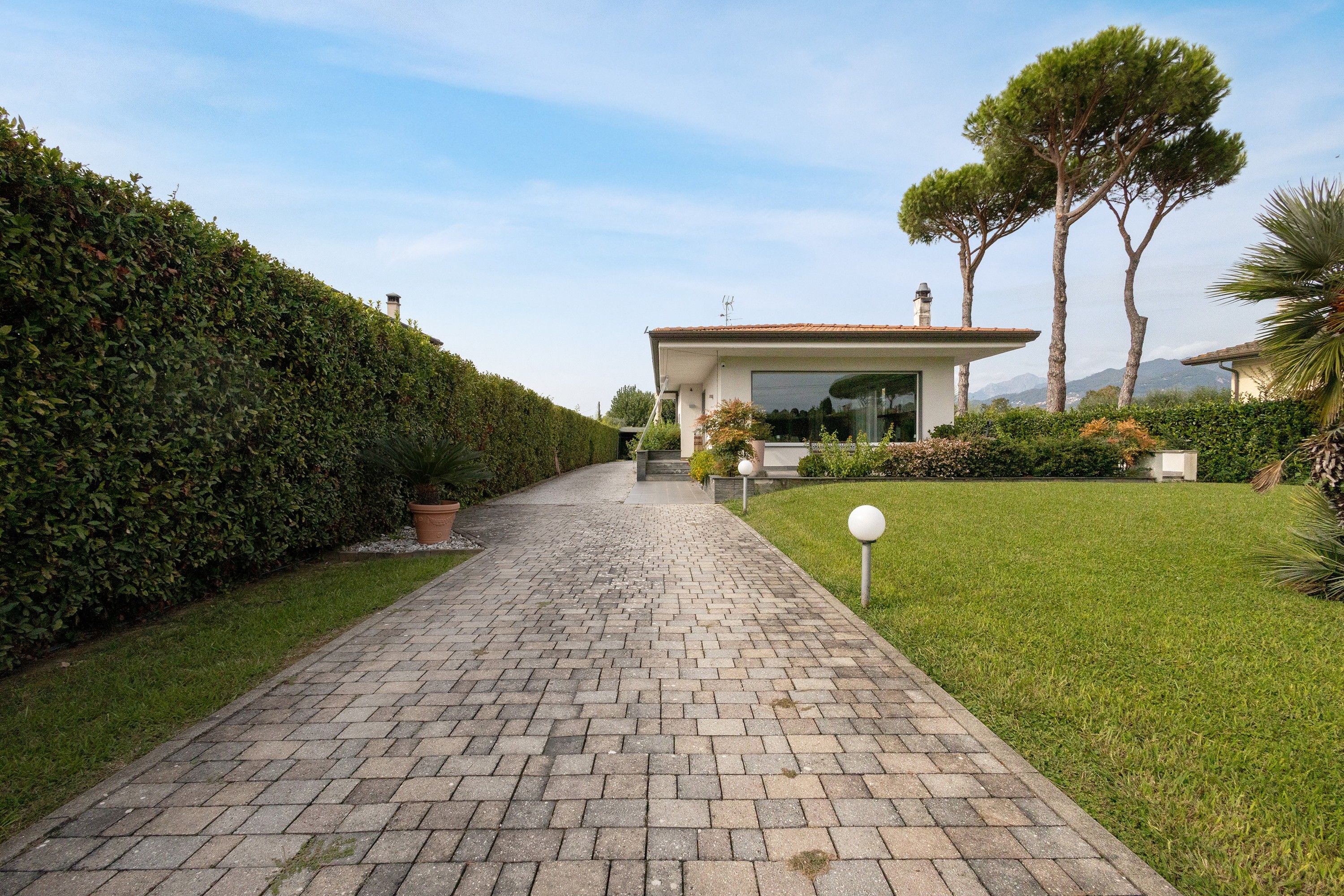 Un chemin pavé mène à une maison moderne dotée d’une grande fenêtre, qui est entourée de haies bien entretenues et d’une pelouse verdoyante.