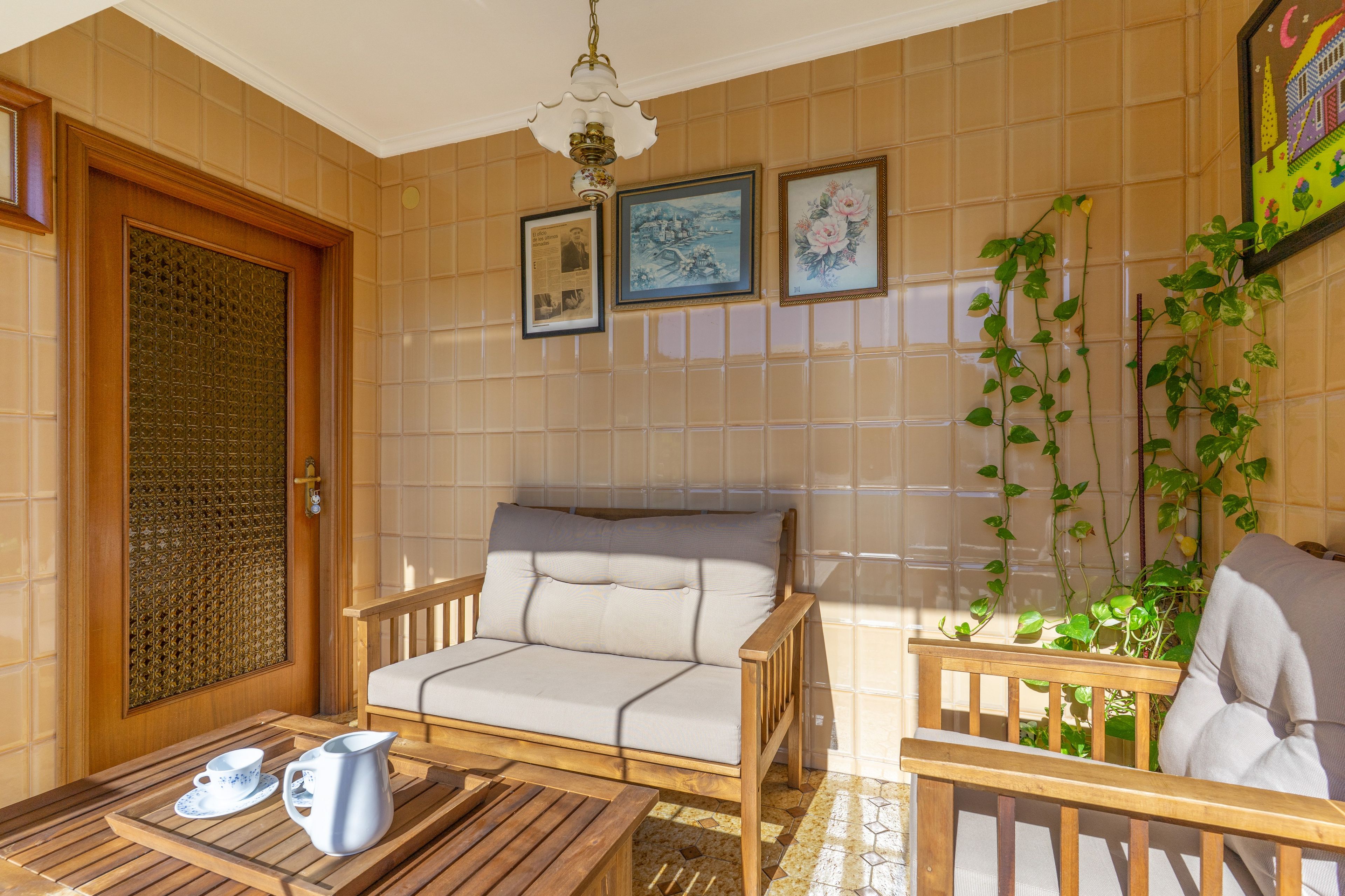 A sunlit room with a wooden door, a cushioned chair, a small table with a pitcher and cups, and a wall with framed pictures and a climbing plant.