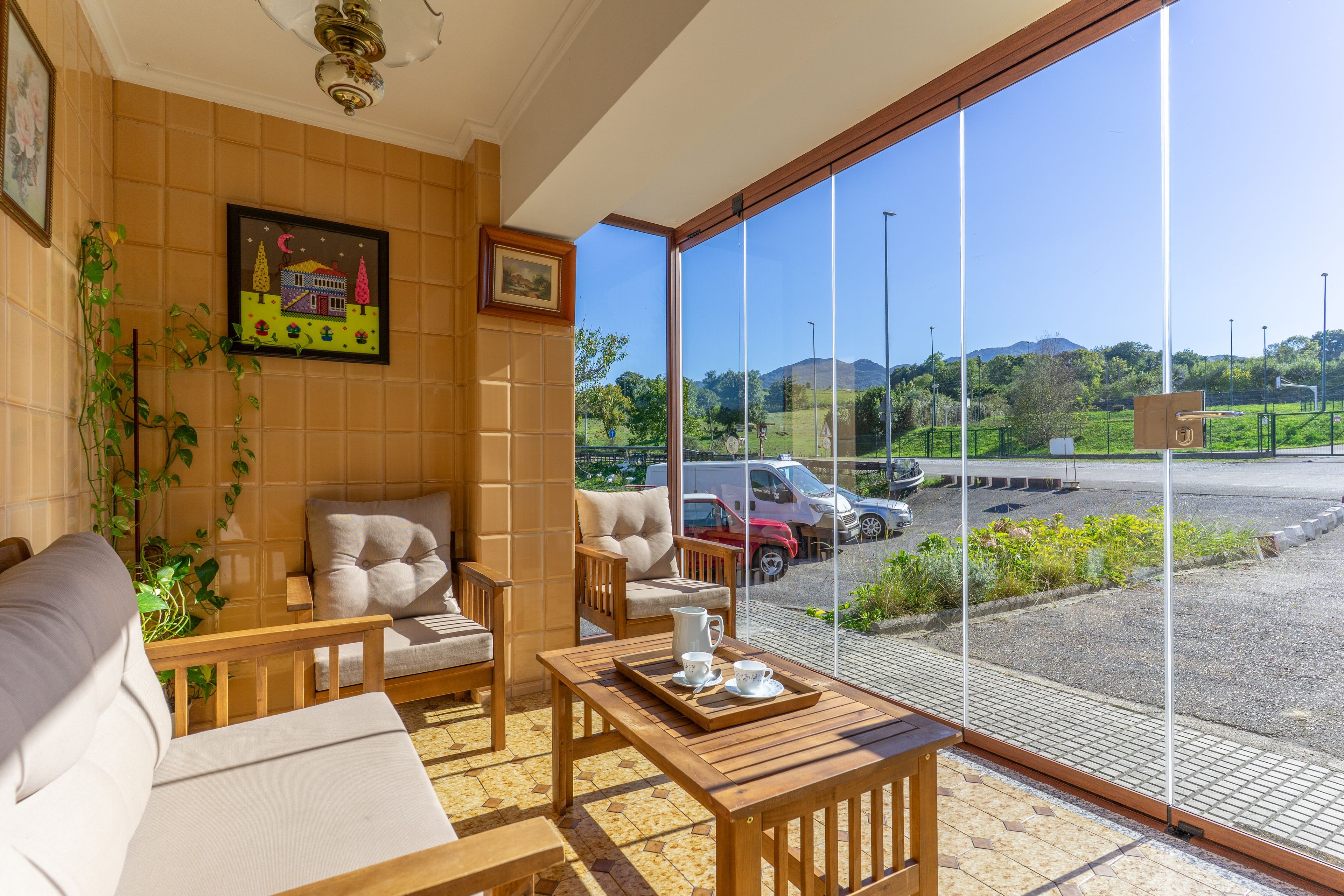 En balkon med et træbord dækket til the, en sofa og en stol, med udsigt over en parkeringsplads med biler og et bjerglandskab.