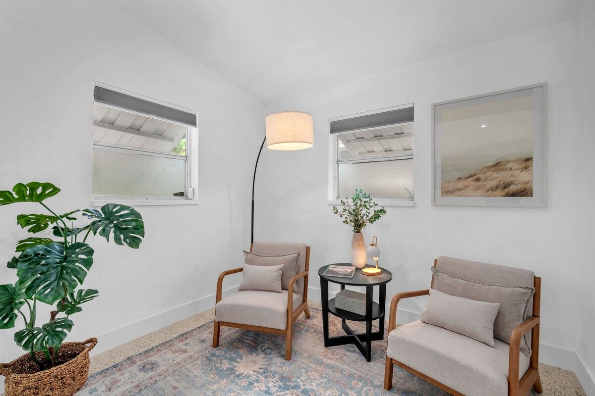 A room with two wooden chairs, a small table with a lamp, a potted plant, and artwork on the walls.