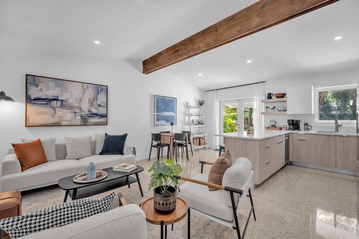 Ein modernes Wohnzimmer mit Sofa, Couchtisch und Sesseln, ein Essbereich mit Tisch und Stühlen sowie eine Küche mit Schränken und einem Fenster.