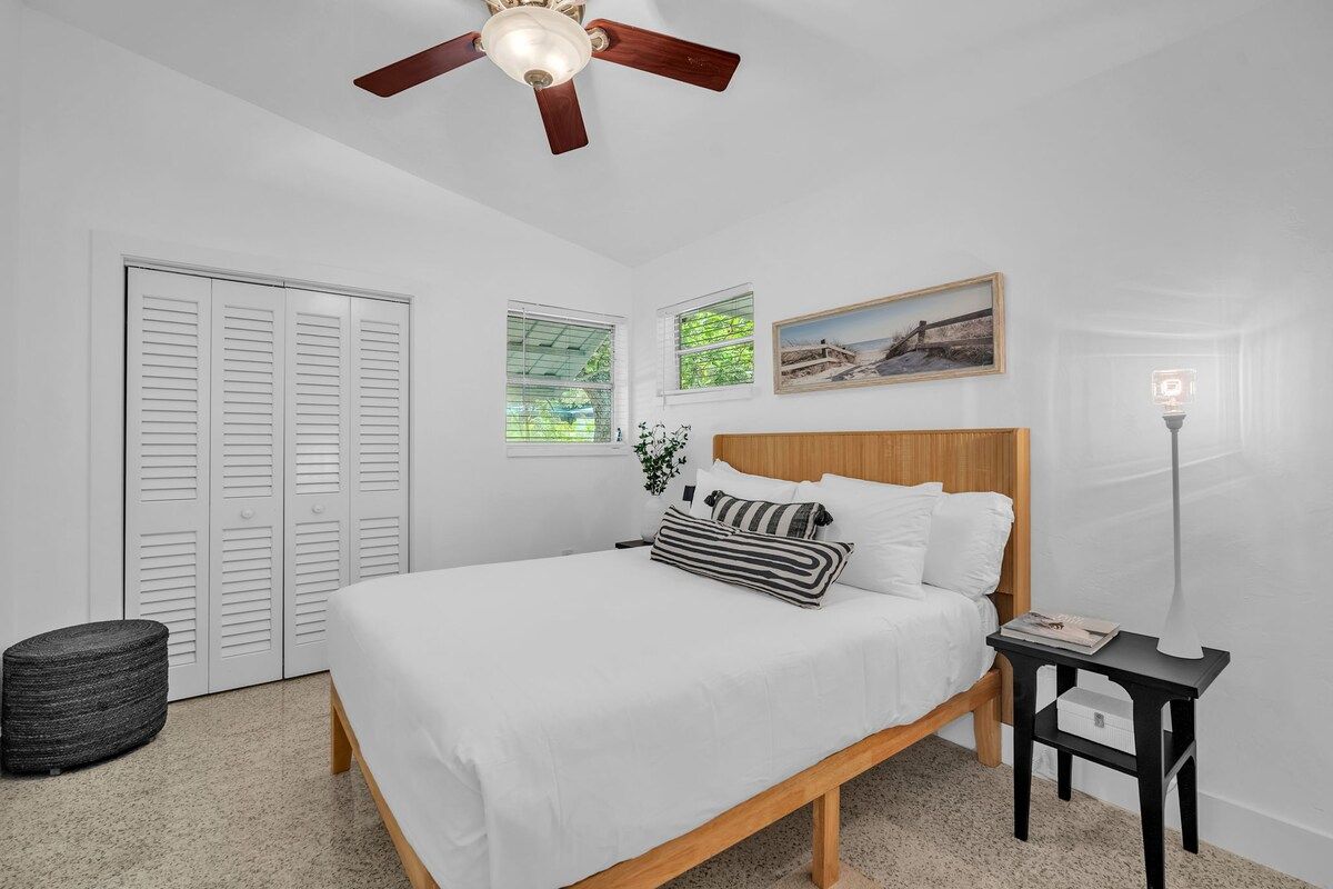 Ein Schlafzimmer mit Bett, Deckenventilator, Nachttisch, Lampe, Fenster und Tür.