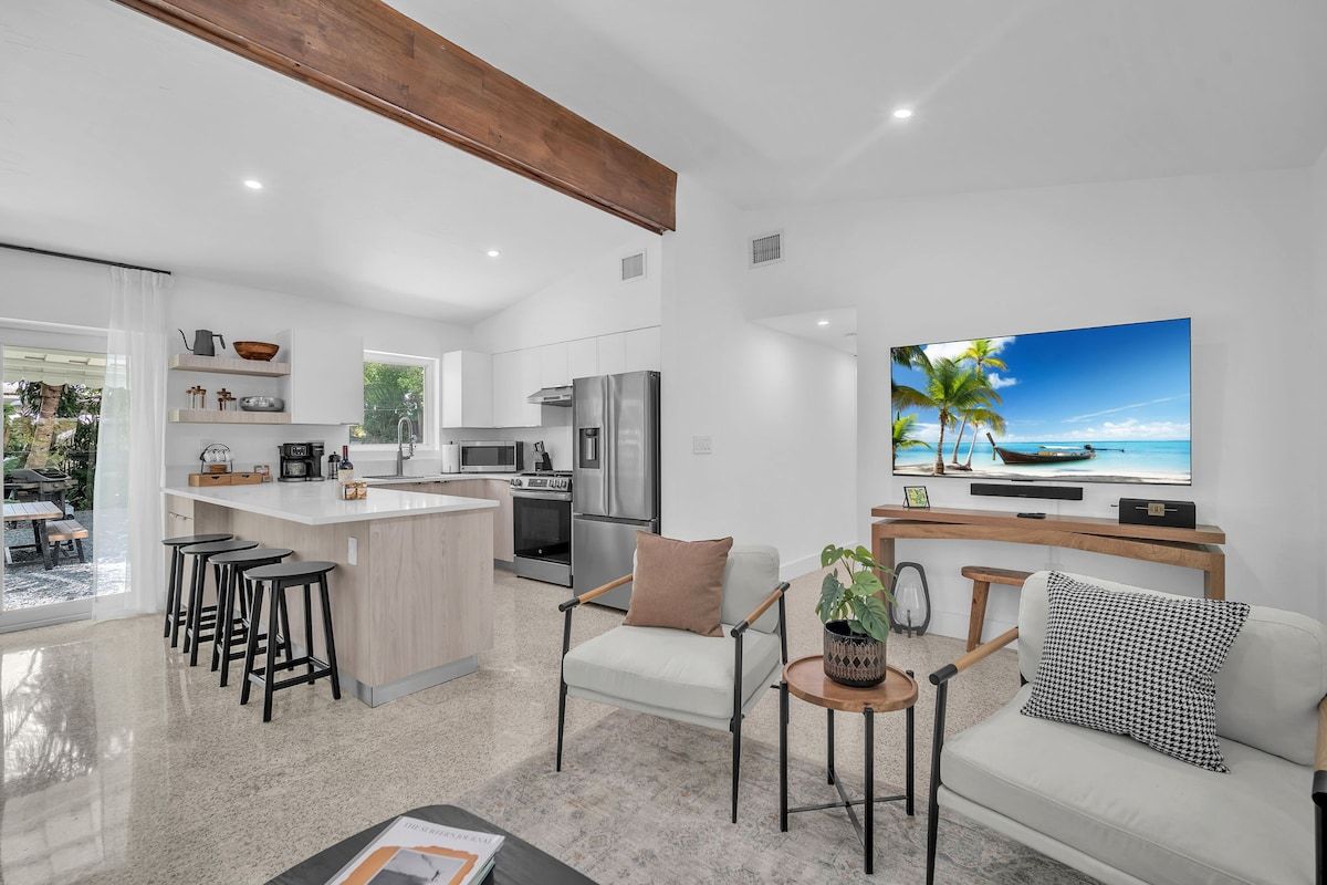 A modern kitchen with a central island, a dining area with bar stools, a seating area with a sofa and armchairs, and a large flat-screen TV mounted on the wall.