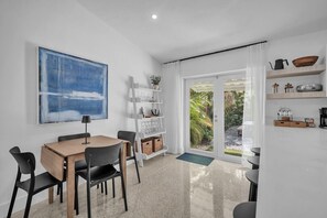 Ein moderner Essbereich mit Holztisch und Stühlen, einer weißen Wand mit einem großen abstrakten Gemälde und Blick auf Grün durch eine Glastür.