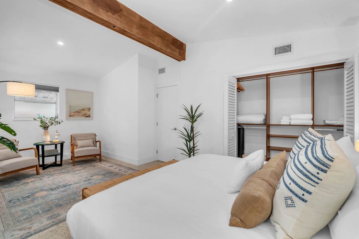Ein Schlafzimmer mit einem großen Bett, einem Kleiderschrank, einem kleinen Tisch mit Lampe, zwei Stühlen und einer Pflanze.
