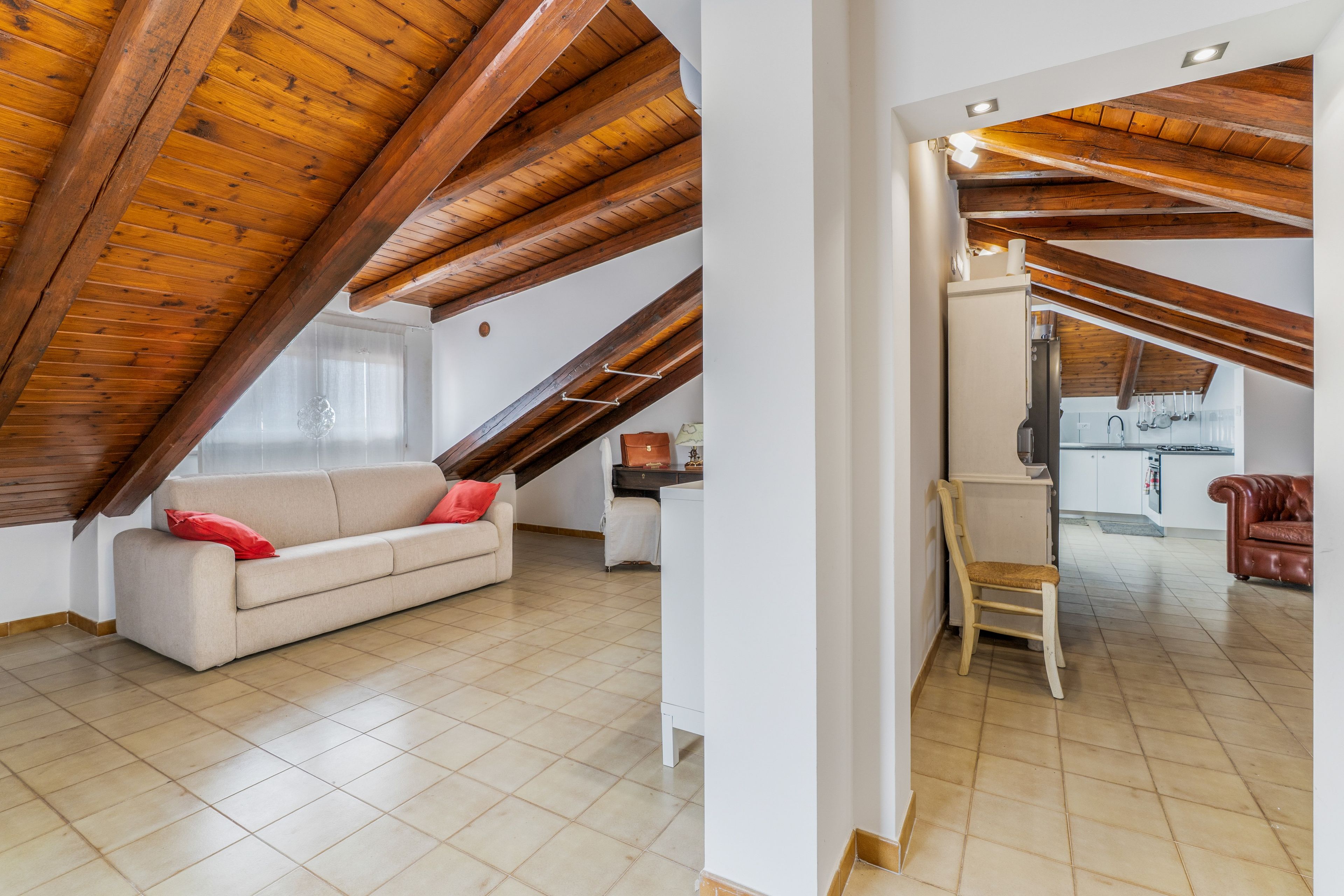 Un'ampia stanza mansardata con soffitto in legno a vista, un divano beige con cuscini rossi, una sedia in legno e un angolo cottura visibile attraverso una porta aperta.