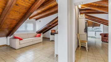 Ein geräumiges Dachzimmer mit einer hohen Holzbalkendecke, ein beiges Sofa mit roten Kissen, ein Holzstuhl und ein durch einen offenen Durchgang sichtbarer Küchenbereich.