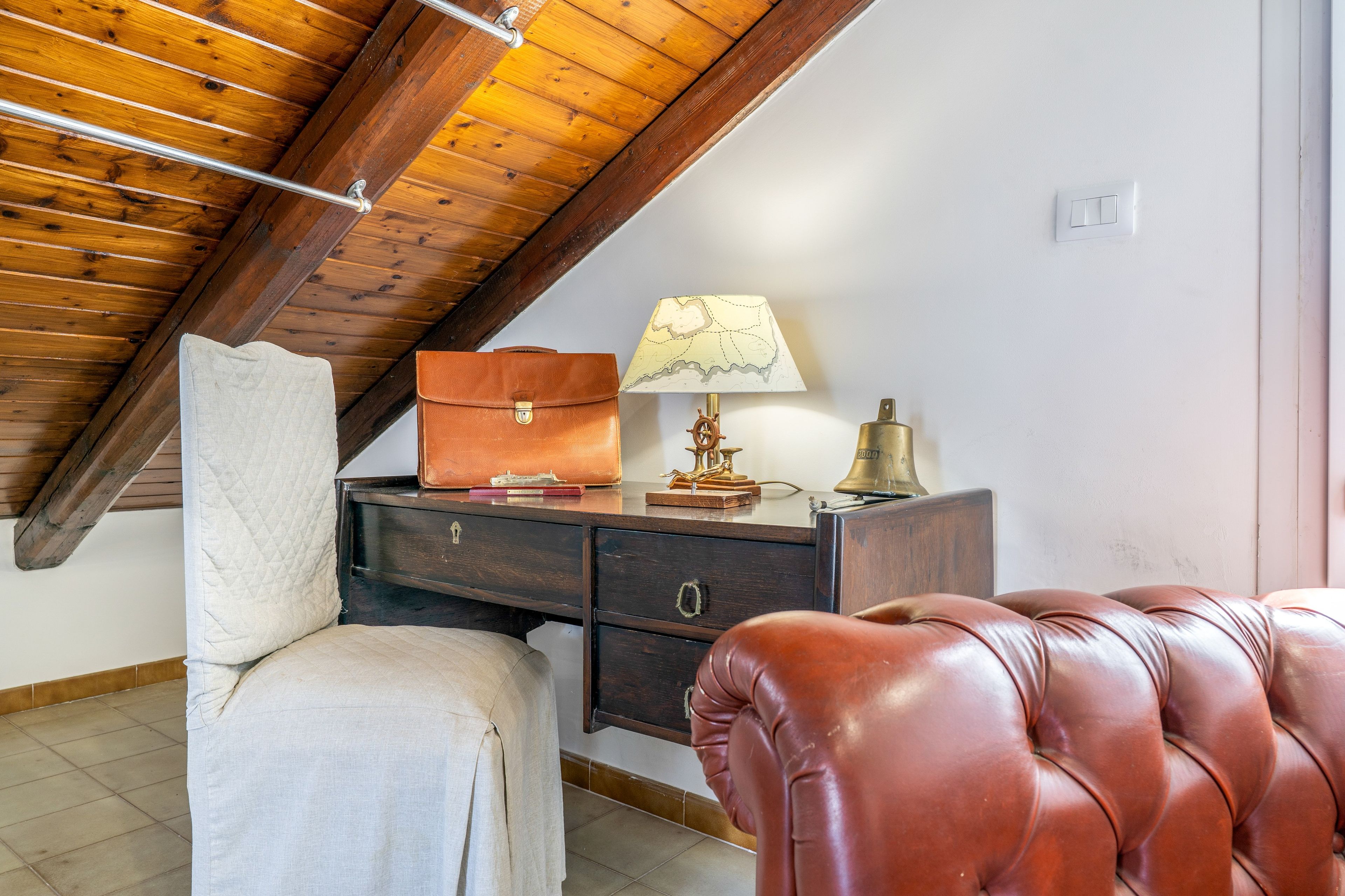 Ein Zimmer mit schrägem Dach, einem Schreibtisch mit Lampe, einem Ledersessel und einer gepolsterten Bank.