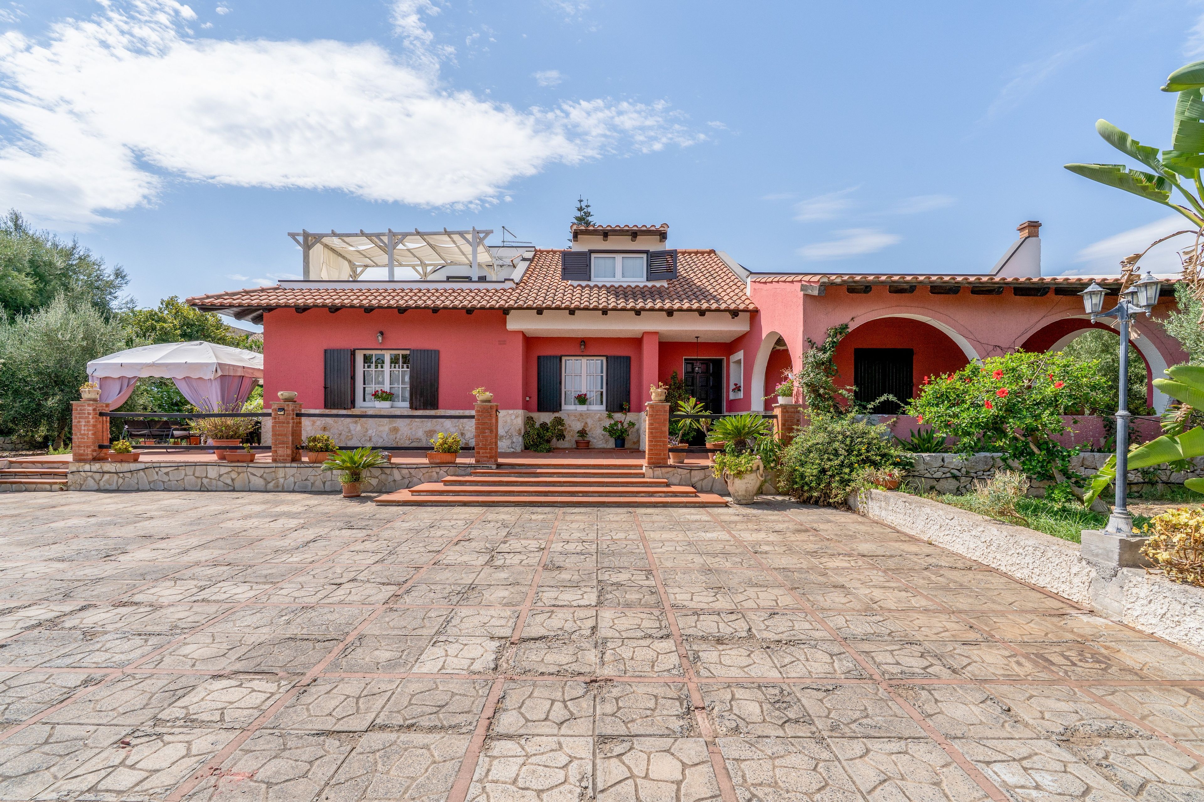 Une villa rouge avec un toit en tuiles, une terrasse en pierre et un jardin agrémenté de plantes en pot.