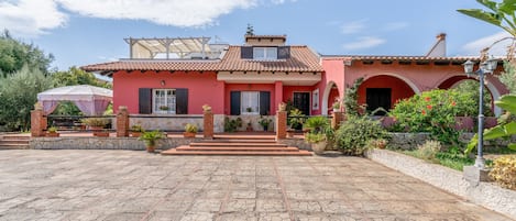 A red villa with a tiled roof, a stone patio, and a garden with potted plants.