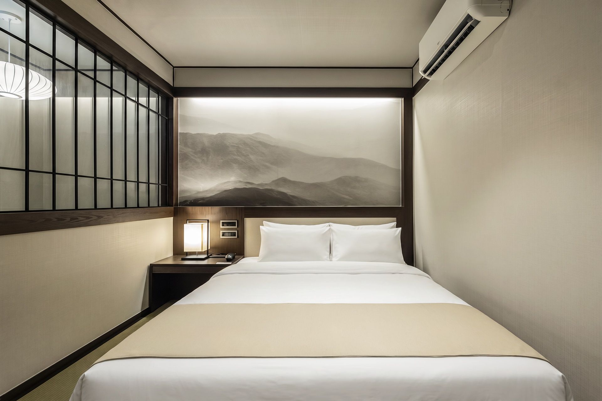 Quarto de hotel com cama, mesa de cabeceira, luminária e ar-condicionado fixado na parede.