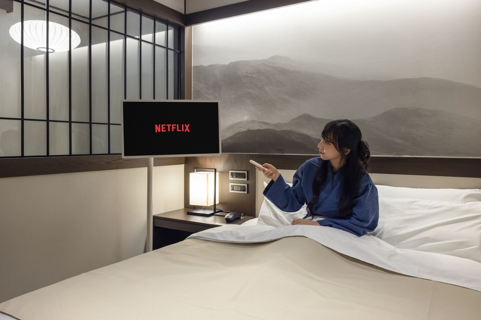 Una habitación con una cama, una mesita de noche con una lámpara, un televisor montado en la pared mostrando 'NETFLIX' y vistas a la montaña a través de una ventana.