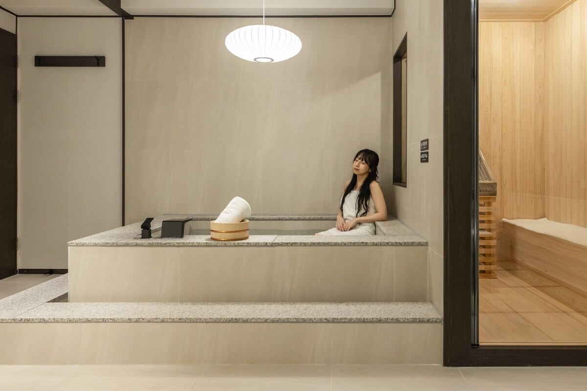 A woman sitting on a bench in a room with a wooden floor, a stone bench, a towel, and a small table with a towel and a black object.