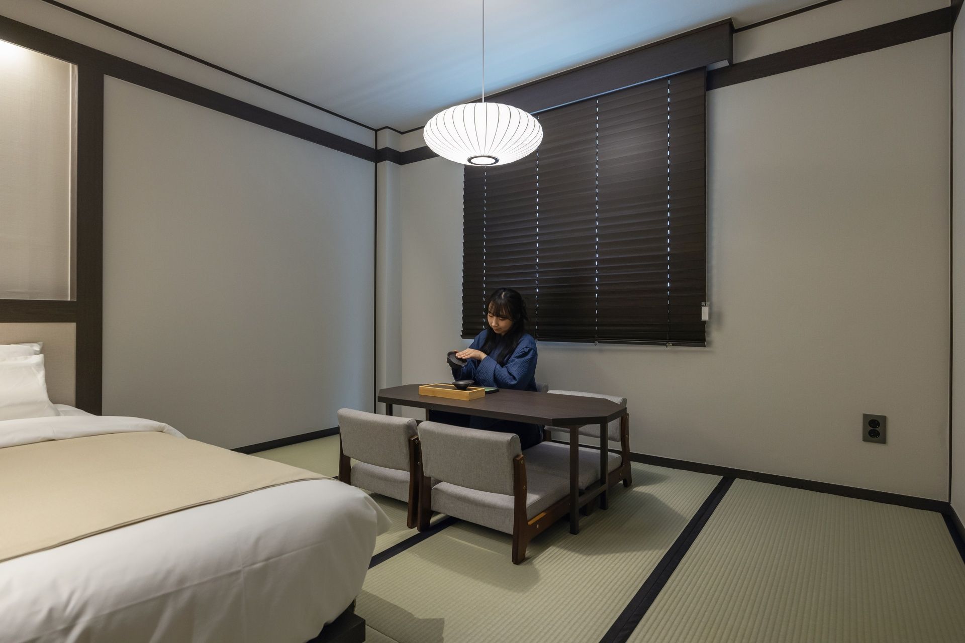 Habitación de hotel con cama, una mesa con sillas, ventana con persianas y lámpara colgante.