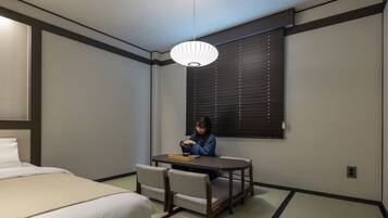 Habitación de hotel con cama, una mesa con sillas, ventana con persianas y lámpara colgante.