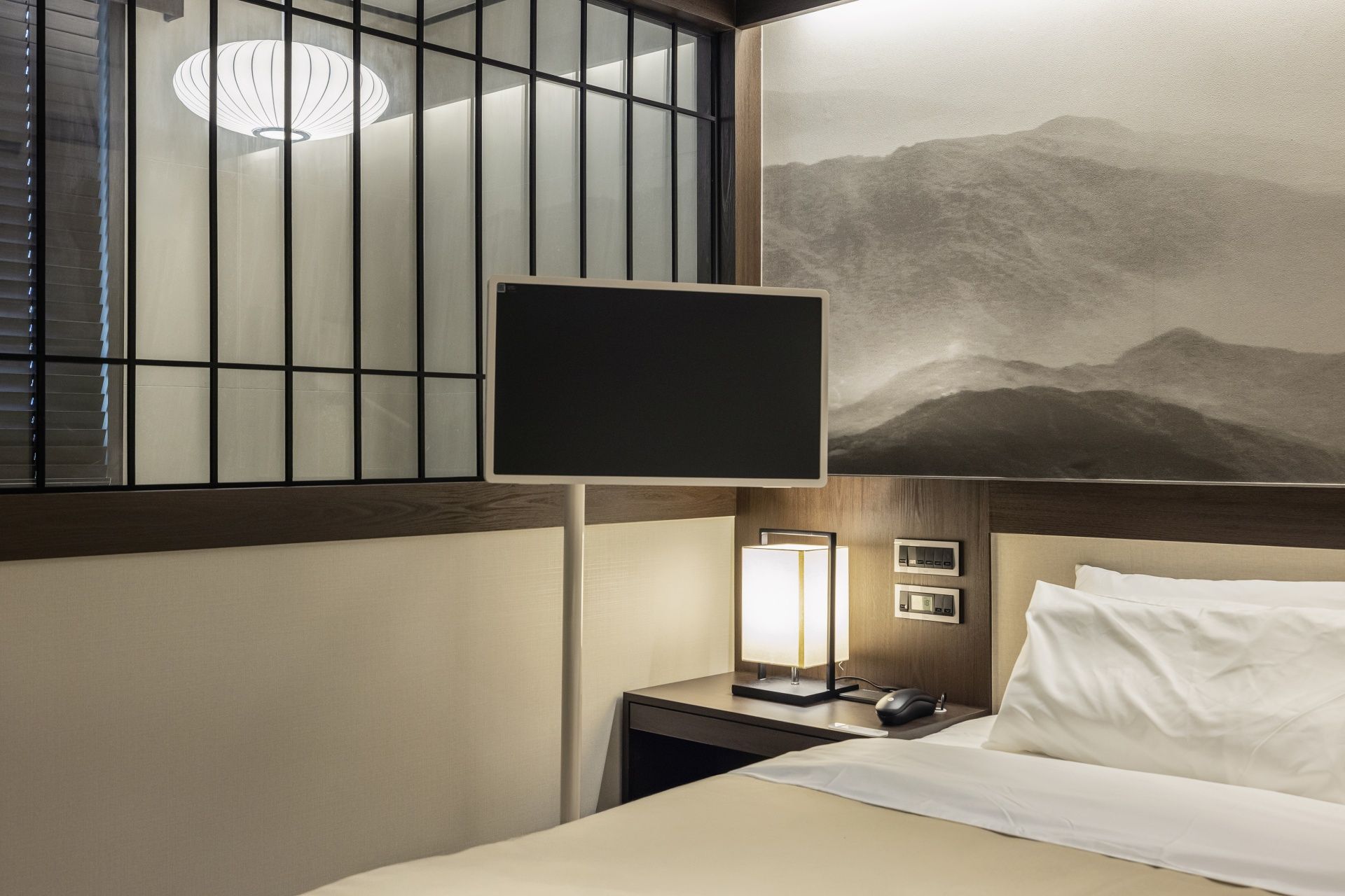 A hotel room with a bed, a nightstand, a wall-mounted TV, and a painting of mountains.