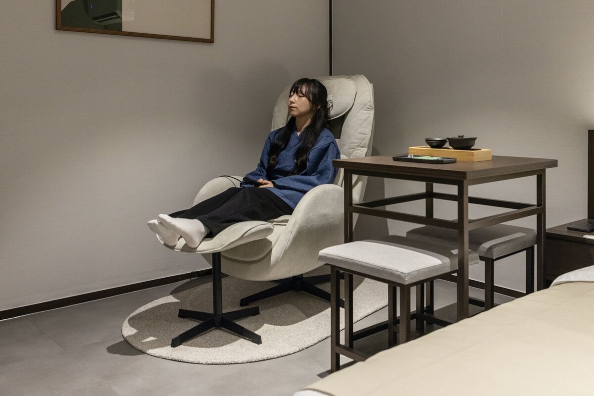 A room with a reclining chair, a small table with a bowl and a cup, and a bed.