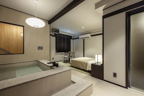 A modern hotel room with a bathtub, a bed, a desk, and a window with blinds.