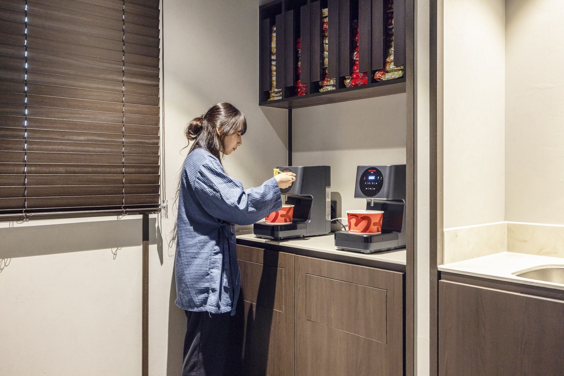 A person using a coffee machine in a room with a window, a shelf with items, and a sink.