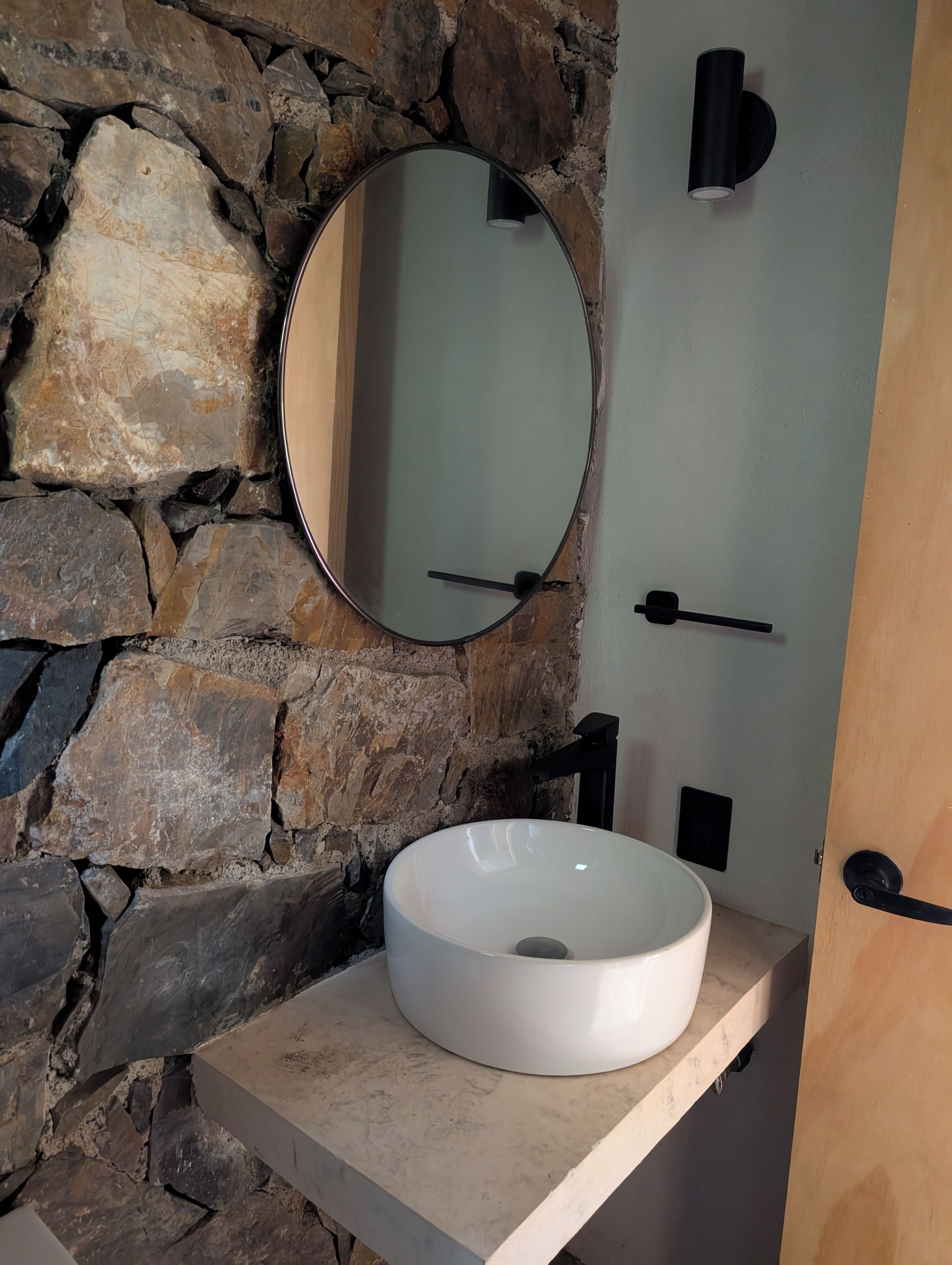 A bathroom with a stone wall, a round mirror, a white sink, and a door with a black handle.