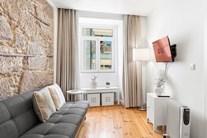 A modern living room with a stone wall, a grey sofa, a window with curtains, a small table with a lamp, and a radiator. - GuestReady - Cozy Retreat in Porto Center (Porto)