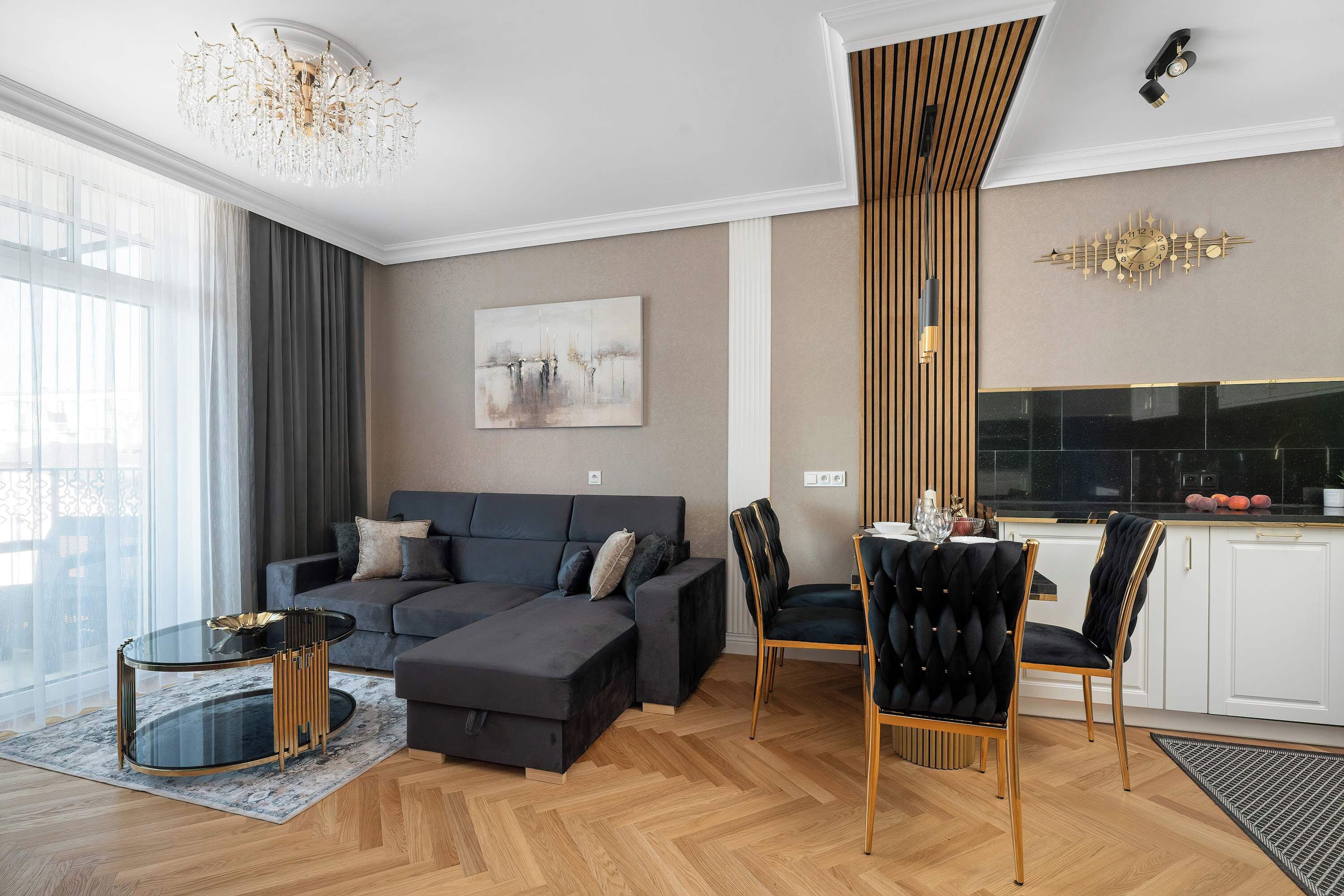 Ein modernes Wohnzimmer mit einer Couch, einem Hocker und Sesseln, ein Essbereich mit Tisch und Stühlen sowie eine Küche mit schwarzem Arbeitsplatz.