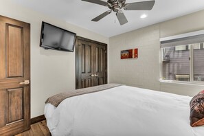 Ein Schlafzimmer mit einem Bett, einem Deckenventilator, einem an der Wand montierten Fernseher und einem Fenster mit Ausblick.