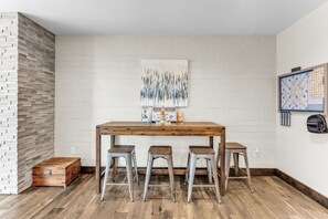 Ein Zimmer mit einem Holztisch, drei Metallhockern, einer weißen Ziegelwand, einem großen abstrakten Gemälde und einem Scrabble-Spiel.